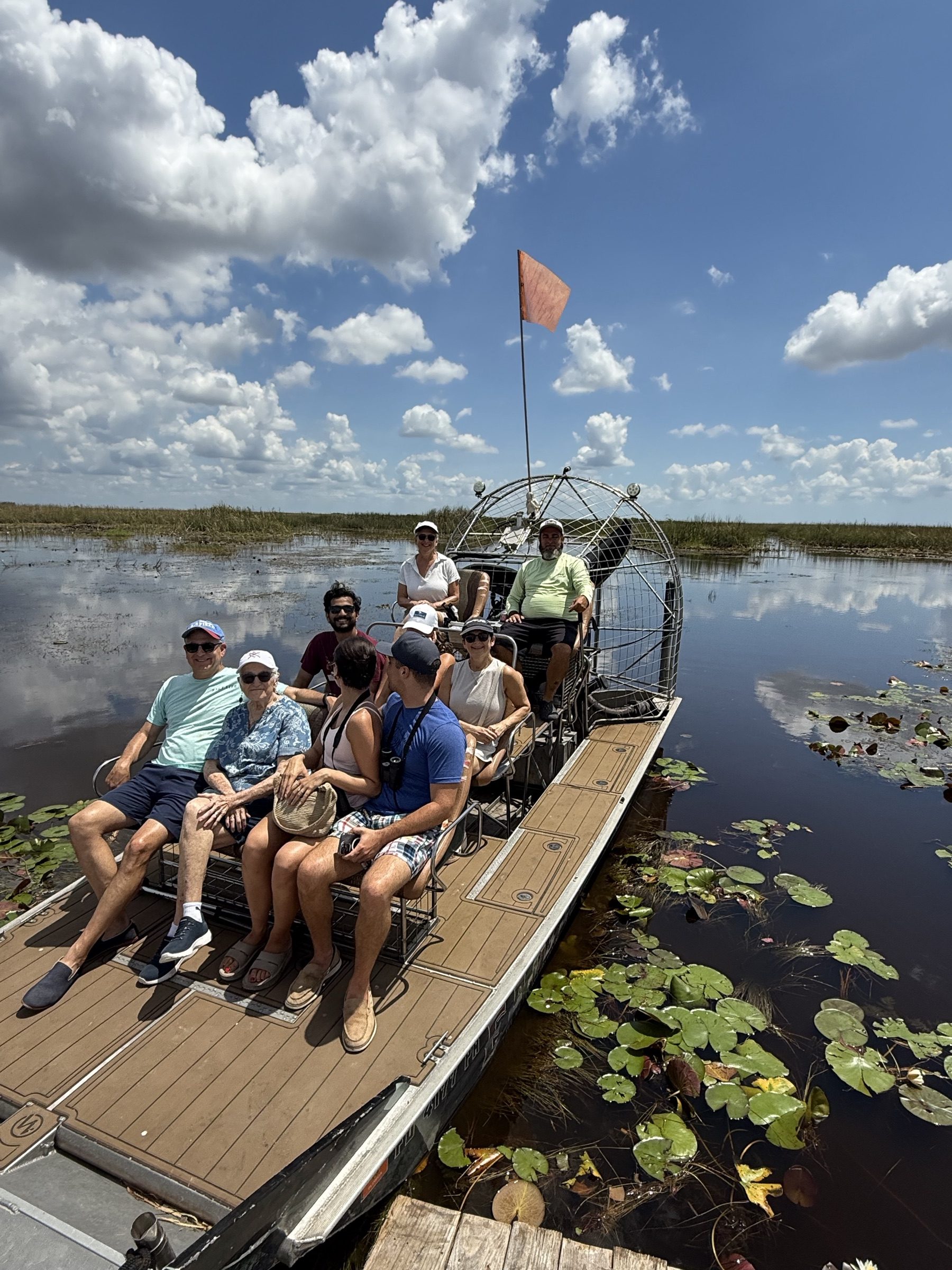 A full airboat of guests gliding over glassy Everglades water reflecting the clouds